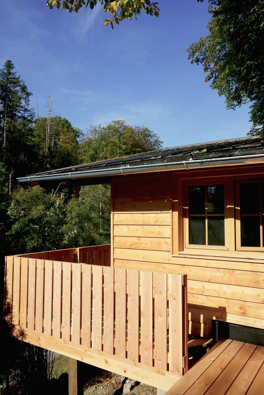 Ecke der renovierten Holzhütte mit Balkon, dahinter Fichten und blauer Himmel